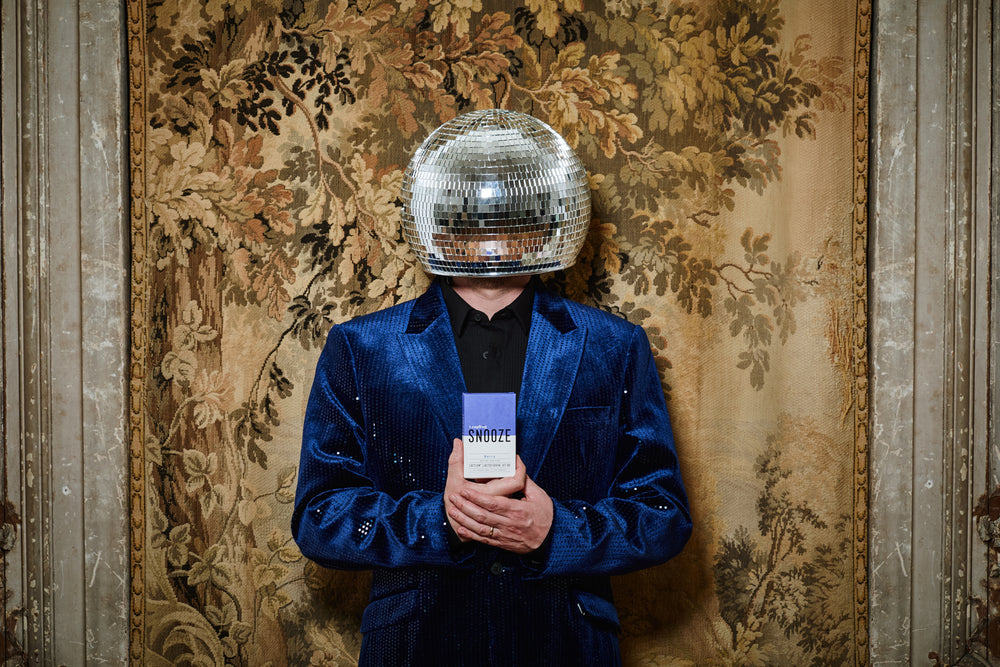 Person wearing a blue jacket and disco ball helmet against an ornate wall holding Leapfrog SNOOZE a natural supplement sleep aid.